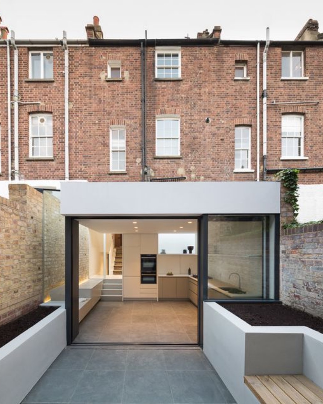 White flat-roofed rear extension with sliding doors, raised planters and a bench